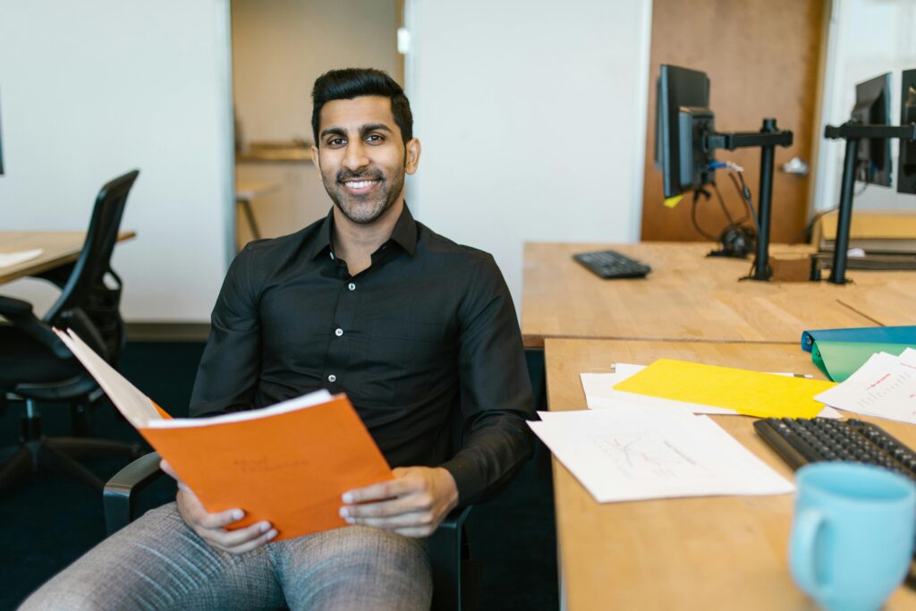 pexels photo 7581004 7581004 Smiling businessman holding documents at his desk, embracing productivity in a modern office.